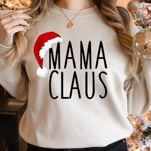 May include: A cream-colored sweatshirt with the text "MAMA CLAUS" in black lettering. A red and white Santa hat is to the left of the text.