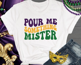May include: White t-shirt with the text "POUR ME SOMETHING MISTER" in green, yellow, and purple letters. The shirt is on a wooden surface with a gold fleur-de-lis and a pair of blue jeans with a gold butterfly mask.