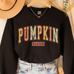 Może przedstawiać: Czarna bluza z nadrukiem w stylu vintage, na kt&oacute;rym widnieje napis "Pumpkin Season" w kolorowych literach.