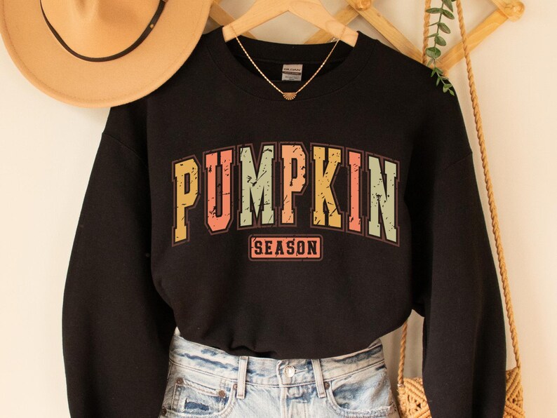 Może przedstawiać: Czarna bluza z nadrukiem w stylu vintage, na kt&oacute;rym widnieje napis "Pumpkin Season" w kolorowych literach.