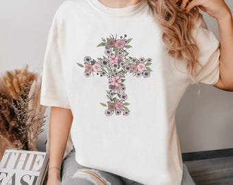 May include: Cream-colored t-shirt featuring a floral cross design. The cross is composed of pink, purple, and green flowers and leaves. The shirt has short sleeves and a relaxed fit, suitable for casual wear.