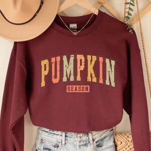 Może przedstawiać: Bordowy sweter z grafiką w stylu vintage, na kt&oacute;rej widnieje napis "Pumpkin Season" w kolorze pomarańczowym, ż&oacute;łtym i zielonym.