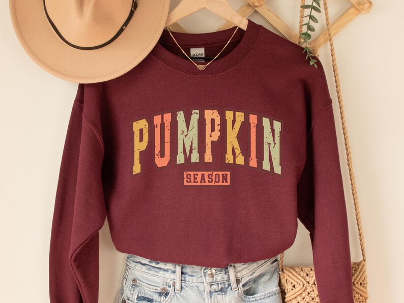 Może przedstawiać: Bordowy sweter z grafiką w stylu vintage, na kt&oacute;rej widnieje napis "Pumpkin Season" w kolorze pomarańczowym, ż&oacute;łtym i zielonym.