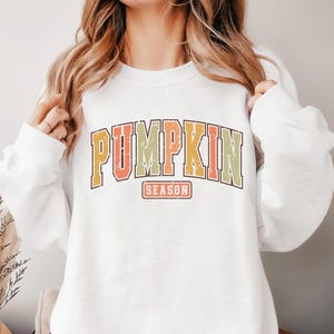 Może przedstawiać: Biała bluza z nadrukiem w stylu vintage, na kt&oacute;rym widnieje napis "Pumpkin Season" w kolorze pomarańczowym, ż&oacute;łtym i zielonym.
