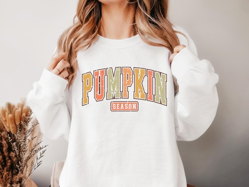 Może przedstawiać: Biała bluza z nadrukiem w stylu vintage, na kt&oacute;rym widnieje napis "Pumpkin Season" w kolorze pomarańczowym, ż&oacute;łtym i zielonym.