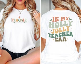 May include: White t-shirt with the word "TEACHER" in a colorful, retro font on the front. The back of the shirt reads "IN MY HOLLY JOLLY TEACHER ERA" in a similar style. The design includes a Christmas tree graphic.