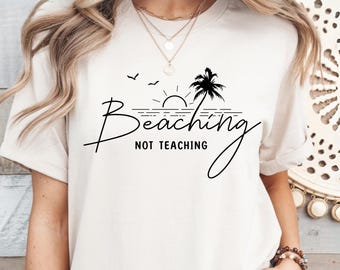 May include: White t-shirt with a black graphic design of a palm tree, sun, and ocean waves. The text "Beaching Not Teaching" is written in a cursive font.
