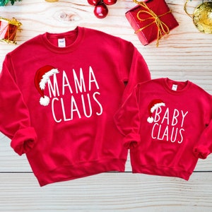 May include: Red Christmas themed sweatshirts with white text. The larger sweatshirt says "Mama Claus" with a Santa hat graphic. The smaller sweatshirt says "Baby Claus" with a Santa hat graphic.