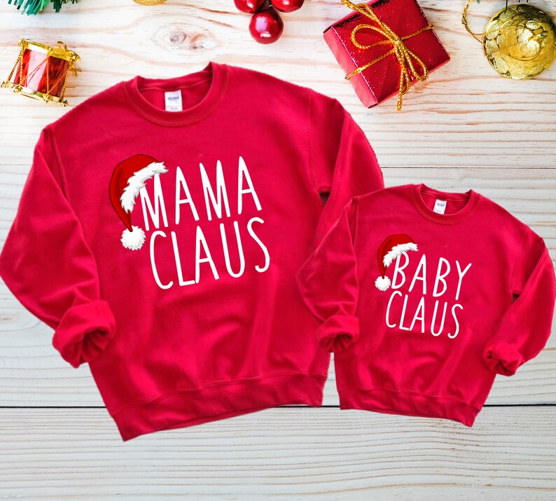 May include: Red Christmas themed sweatshirts with white text. The larger sweatshirt says "Mama Claus" with a Santa hat graphic. The smaller sweatshirt says "Baby Claus" with a Santa hat graphic.