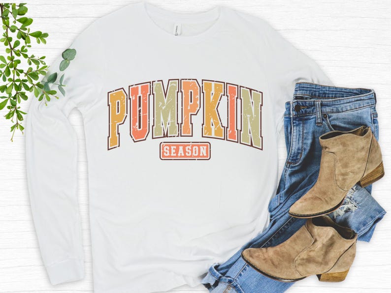 Może przedstawiać: Biała koszula z długim rękawem z grafiką w stylu vintage, kt&oacute;ra głosi "Pumpkin Season" w kolorze pomarańczowym, ż&oacute;łtym i czerwonym.