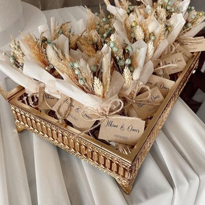 Op de afbeelding: Een gouden dienblad gevuld met kleine boeketten gedroogde bloemen, verpakt in bruin papier. Elk boeket heeft een klein kaartje met de tekst "Ilkim & Omer" en een datum.