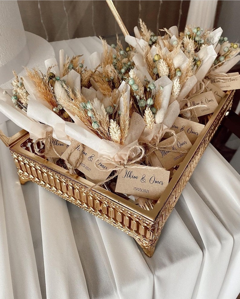 Op de afbeelding: Een gouden dienblad gevuld met kleine boeketten gedroogde bloemen, verpakt in bruin papier. Elk boeket heeft een klein kaartje met de tekst "Ilkim & Omer" en een datum.