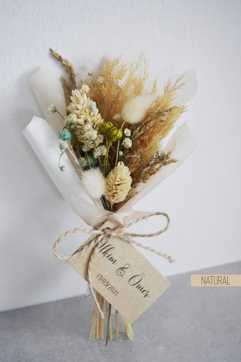 Op de afbeelding: Een klein boeket gedroogde bloemen, verpakt in wit tissuepapier en vastgebonden met touw. Het boeket bevat gedroogd pampasgras, konijnenstaarten en andere gedroogde bloemen in beige, wit en groene tinten. Een bruin papieren label is bevestigd aan het boeket met de tekst "Ilkim & &Ouml;mer 13/05/2021" en het woord "NATUURLIJK".
