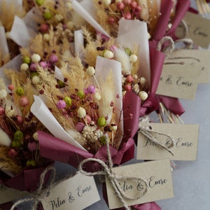 Op de afbeelding: Een verzameling kleine boeketten gedroogde bloemen, verpakt in bordeauxrood papier met een bruin label waarop "Peln & Emre" staat.