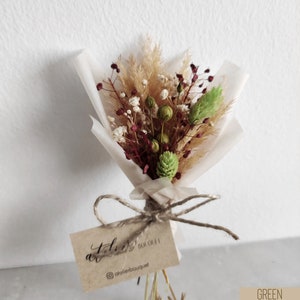 Op de afbeelding: Een klein boeket gedroogde bloemen in tinten bruin, groen en bordeauxrood, verpakt in wit tissuepapier en vastgebonden met touw. Een klein bruin kaartje met de tekst "atelier bouquet" is bevestigd.