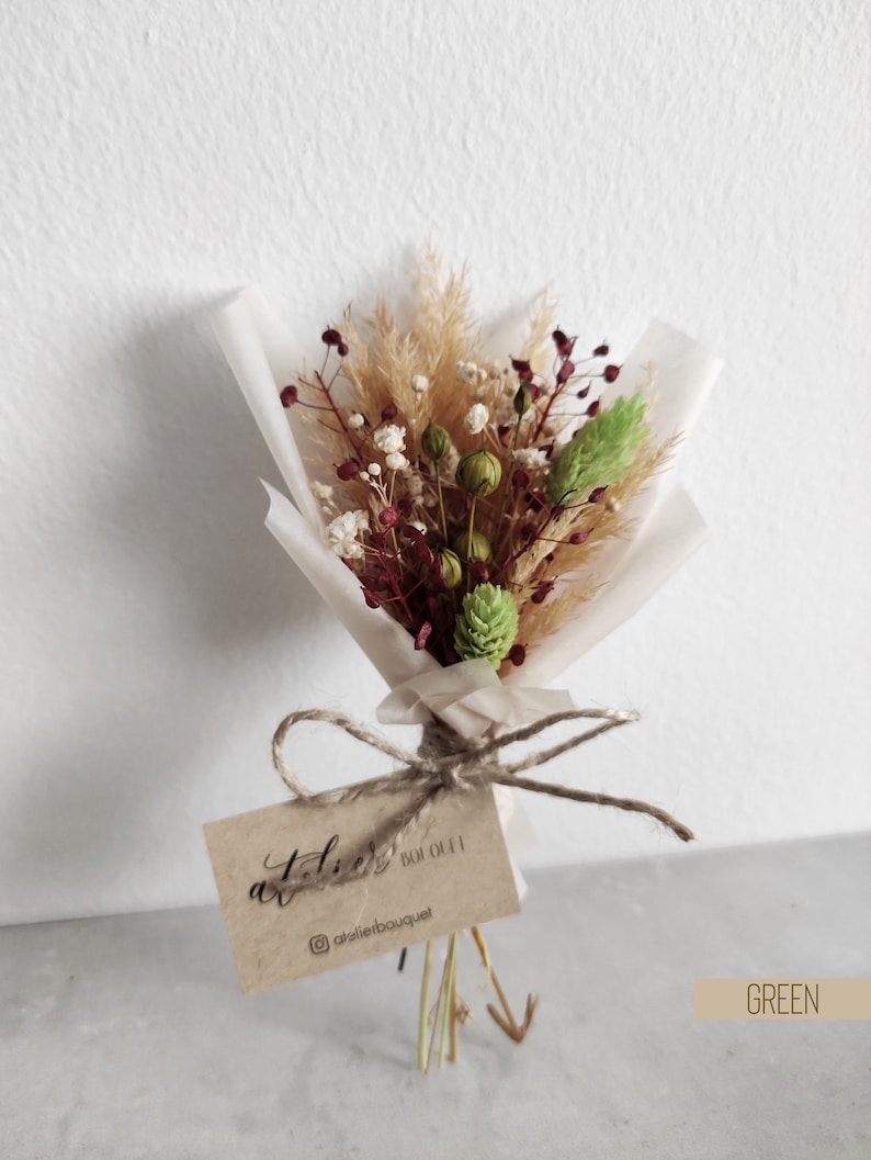 Op de afbeelding: Een klein boeket gedroogde bloemen in tinten bruin, groen en bordeauxrood, verpakt in wit tissuepapier en vastgebonden met touw. Een klein bruin kaartje met de tekst "atelier bouquet" is bevestigd.