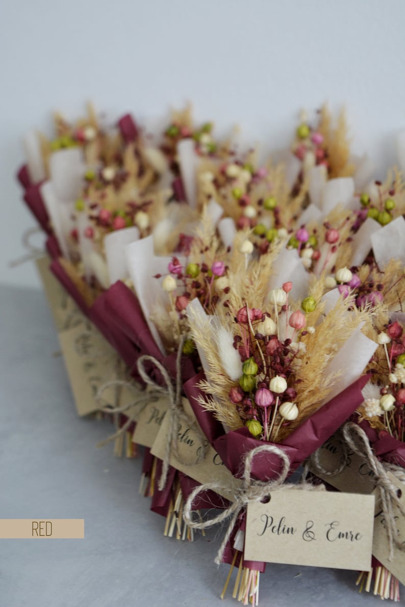 Op de afbeelding: Een groep kleine boeketten verpakt in bordeauxrood papier met een bruin label waarop "Pelin & Emre" staat. De boeketten zijn gemaakt van gedroogde bloemen en grassen in tinten roze, groen en beige.