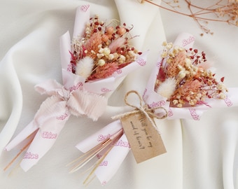 Op de afbeelding: Twee kleine boeketten gedroogde bloemen verpakt in roze papier met de namen "Chloe" en "Barbara" erop gedrukt. Een bruine kaart met de tekst "Wil je mijn bruidsmeisje zijn?" is aan een van de boeketten bevestigd.