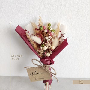 Op de afbeelding: Een klein boeket gedroogde bloemen in tinten roze, wit en groen, verpakt in bordeauxrood papier en vastgebonden met touw. Het boeket is ongeveer 15 tot 17 cm hoog en heeft een label met de tekst "atelier" en een social media-handvat.