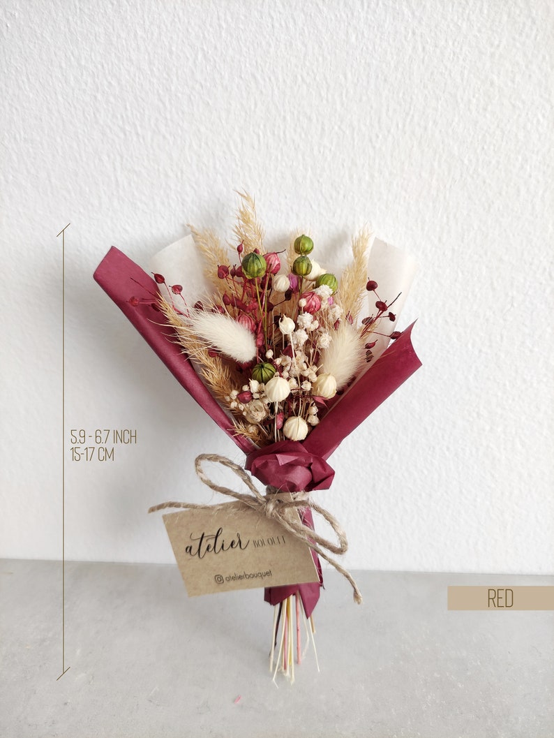 Op de afbeelding: Een klein boeket gedroogde bloemen in tinten roze, wit en groen, verpakt in bordeauxrood papier en vastgebonden met touw. Het boeket is ongeveer 15 tot 17 cm hoog en heeft een label met de tekst "atelier" en een social media-handvat.
