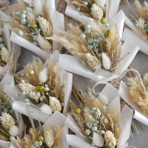 Op de afbeelding: Een verzameling kleine boeketten gedroogde bloemen, verpakt in wit tissuepapier en vastgebonden met touw. De boeketten bevatten gedroogd pampasgras, konijnenstaarten en andere gedroogde bloemen in beige, wit en lichtblauwe tinten.