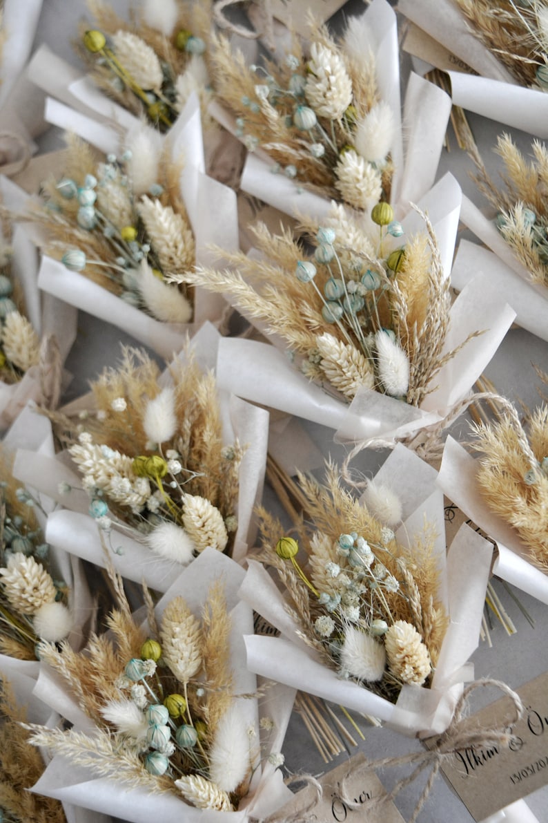 Op de afbeelding: Een verzameling kleine boeketten gedroogde bloemen, verpakt in wit tissuepapier en vastgebonden met touw. De boeketten bevatten gedroogd pampasgras, konijnenstaarten en andere gedroogde bloemen in beige, wit en lichtblauwe tinten.