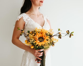 Pode incluir: Uma noiva segura um buquê de girassóis e flores amarelas com caules verdes e bagas marrons. O buquê está envolto em um tecido verde escuro.