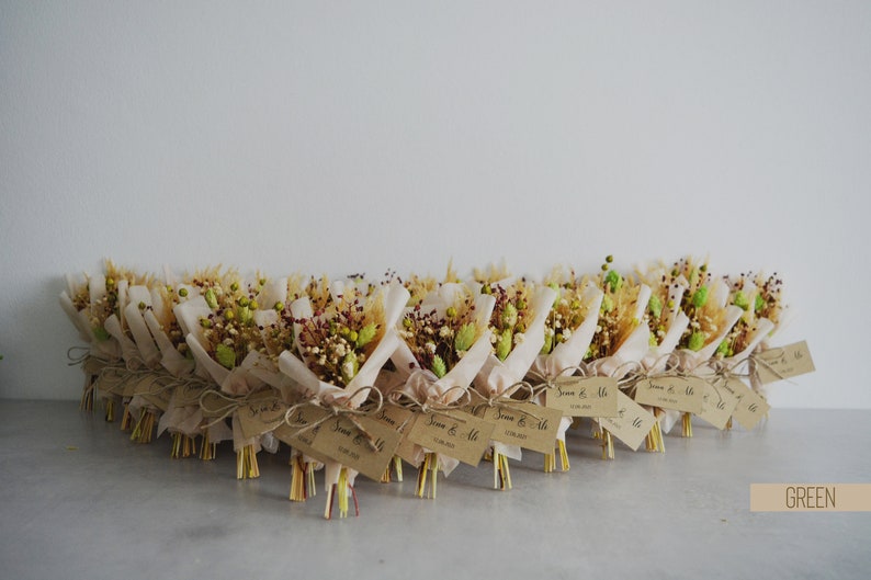 Op de afbeelding: Een rij kleine, rustieke boeketten verpakt in bruin papier met touw en een label met de tekst "Jenna & Mike". De boeketten zijn gemaakt van gedroogde bloemen en groen in tinten bruin, beige en groen.