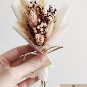 Op de afbeelding: Een klein boeket gedroogde bloemen, verpakt in wit tissuepapier en vastgebonden met touw. Het boeket bevat gedroogd pampasgras, bordeauxrode en witte bloemen en een klein kaartje met de tekst "@atelierbouquet". Het kaartje is vastgebonden aan het touw. Het boeket is gelabeld met "PINK".