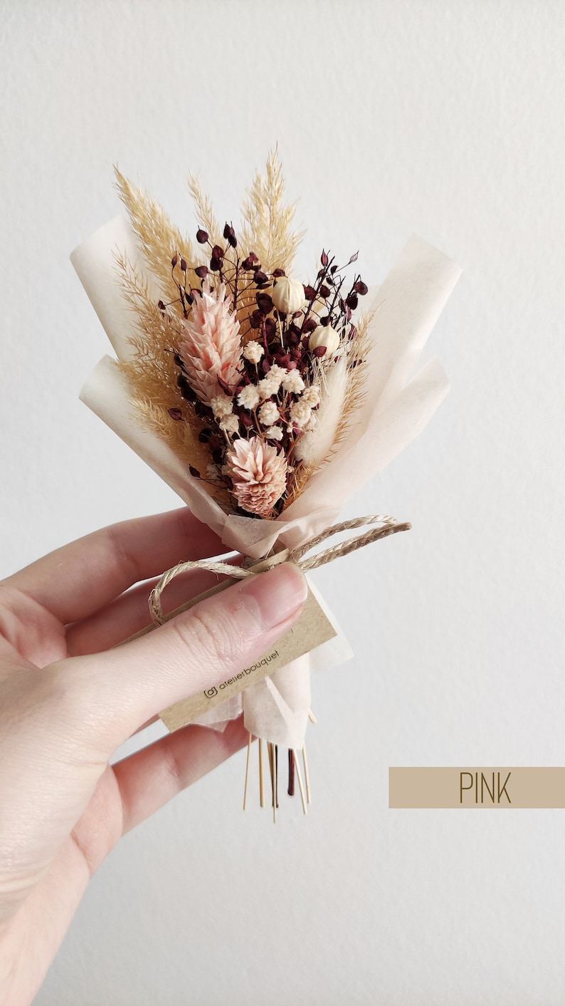 Op de afbeelding: Een klein boeket gedroogde bloemen, verpakt in wit tissuepapier en vastgebonden met touw. Het boeket bevat gedroogd pampasgras, bordeauxrode en witte bloemen en een klein kaartje met de tekst "@atelierbouquet". Het kaartje is vastgebonden aan het touw. Het boeket is gelabeld met "PINK".