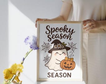 Puede incluir: Un estampado enmarcado con las palabras "Spooky season" y un fantasma de dibujos animados con un sombrero de bruja y una calabaza. El fantasma es blanco, el sombrero es marrón y la calabaza es naranja. Hay telarañas a ambos lados del fantasma.