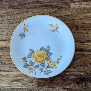 May include: A white ceramic saucer with a gold rim and a floral design featuring yellow roses and green leaves.