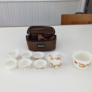 May include: A brown fabric carrying case with a white porcelain tea set. The tea set includes a teapot and six small cups with a gold dragon design.