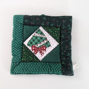 May include: A green quilted Christmas tree pot holder with a red bow. The pot holder is made of different green fabrics with a white and green checkered pattern in the center.