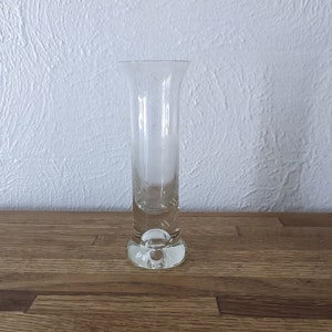 May include: A clear glass bud vase with a rounded base. The vase is empty and sitting on a wooden surface.