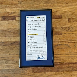 May include: A blue and white framed schedule for the Tampa Bay Rays baseball team. The schedule includes the number of games for each stage of the season, including spring training, regular season, and playoffs. The schedule also includes the names of the teams the Rays will play against. The schedule is signed by the manager.