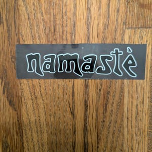 May include: A rectangular black sticker featuring the word namasté written in a stylized white font with a thin light blue outline. The lettering is lowercase and has a distinct, flowing appearance across the center of the sticker surface.