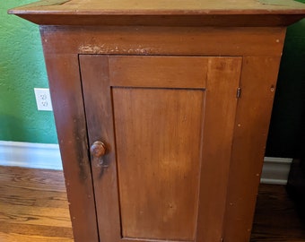 May include: A vintage wooden cabinet with a single door and a simple knob. The cabinet is painted a dark brown color and has a slightly distressed finish.