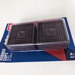 May include: Two brown rubber furniture cups in a clear plastic package. The package is labeled "Rubber Cups" and "Bases de Goma". The cups are 2 inches in diameter.