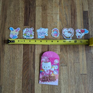 May include: A pink Hello Kitty red envelope with a gold Chinese character and the text "吉祥如意" which translates to "Good Luck and Prosperity". The envelope features a Hello Kitty illustration with a rainbow and a pink heart. There are six Hello Kitty stickers on a wooden surface.