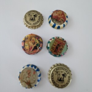 May include: Six vintage bottle caps with various designs. The caps are round and made of metal. They feature images of a dog, a woman, a man, and text. The caps are in various colors, including gold, blue, red, and white.
