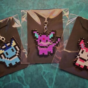 Può includere: Tre portachiavi con ciondoli pixel art in confezioni di plastica. I ciondoli hanno la forma di una creatura con una chiusura argentata. Uno è blu e bianco, uno viola e uno rosa e bianco.