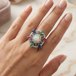 May include: A silver-toned ring featuring a central oval opal surrounded by a halo of small white stones. The design includes radiating accents of pear-shaped blue sapphires, pink rubies, and green emeralds, all set within a circular frame.