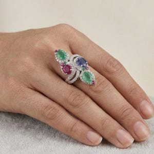 May include: A silver-toned ring featuring a cluster of oval-cut gemstones including two green emeralds, one blue sapphire, and two red rubies, all surrounded by small clear pavé-set stones arranged in a swirling, multi-layered metal band design.