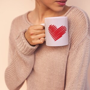 K&ouml;nnte beinhalten: Eine Frau in einem hellrosa Pullover h&auml;lt eine wei&szlig;e Tasse mit einem roten Herzmotiv.
