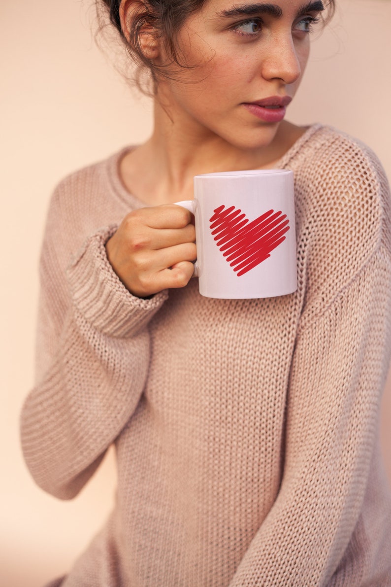 K&ouml;nnte beinhalten: Eine Frau in einem hellrosa Pullover h&auml;lt eine wei&szlig;e Tasse mit einem roten Herzmotiv.