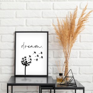 Puede incluir: Un cuadro negro con una impresión en blanco y negro que muestra la palabra "dream" y una ilustración de diente de león. La impresión está sobre una mesa negra, junto a un jarrón con trigo seco y objetos decorativos. El fondo es una pared de ladrillos blancos.