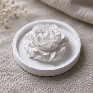 May include: A white circular tray featuring a three-dimensional sculpted flower at its center. The tray has a smooth, matte finish and a raised edge, while the flower displays intricate, layered petals with a realistic, textured appearance.
