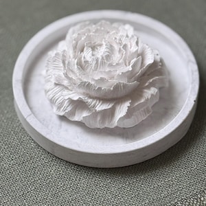 May include: White ceramic flower shaped trinket dish. The dish is round and has a raised flower design in the center. The flower is white and has a textured surface.