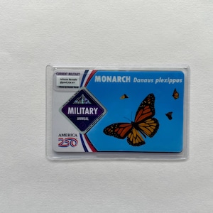 May include: A clear plastic card holder containing a blue and white card. The card features a Monarch butterfly illustration with orange and black wings. Text includes "MONARCH Danaus plexippus" and "MILITARY ANNUAL AMERICA 250".